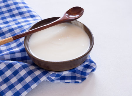 Homemade greek yogurt or sour cream in old clay bowl with wooden spoon and blue checkered fabric or napkin over white wooden background. Food helps the digestive system.の写真素材