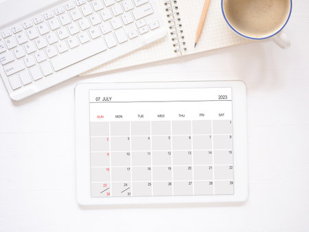 The online calendar app on tablet computer showing screen July 2023, on white workplace and coffee, keyboard, pencil. Reminder with the calendar application for schedule planning. Top view.の写真素材