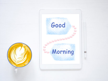 Text "Good Morning" on blue watercolor over digital tablet pc screen, and coffee cup on white wooden background. Positive emotion concept.の写真素材