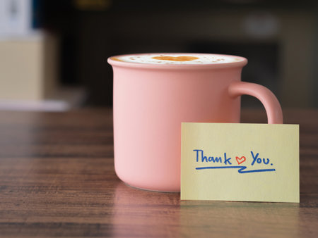 Positive and Thankfulness concept. Handwritten text "thank you" message and hot latte coffee in pink color ceramic mug on brown wooden table.の写真素材