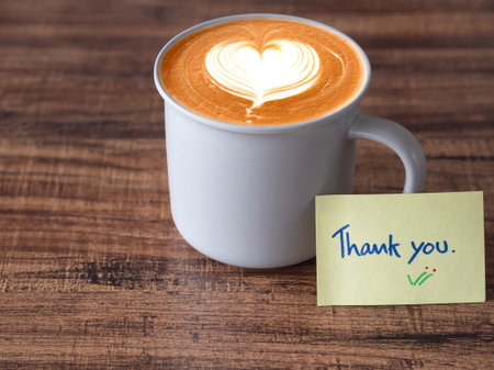 Handwritten text "thank you" message and hot latte coffee in grey color ceramic mug on brown wooden table.の写真素材