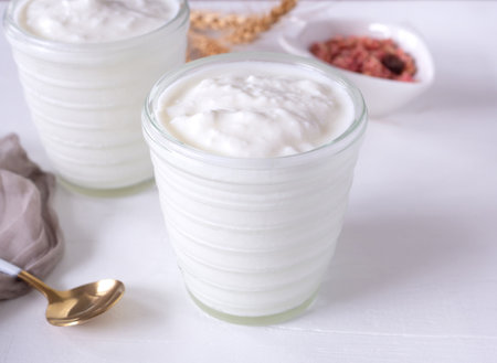 Greek yogurt in cup glass and brown cloth, spoon, granola on white wooden background.の写真素材