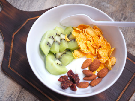 Greek yogurt in a white bowl topped with kiwi, almonds, dried strawberries, and cornflakes, on a wooden cutting board. Top view, flat lay with copy space.の写真素材