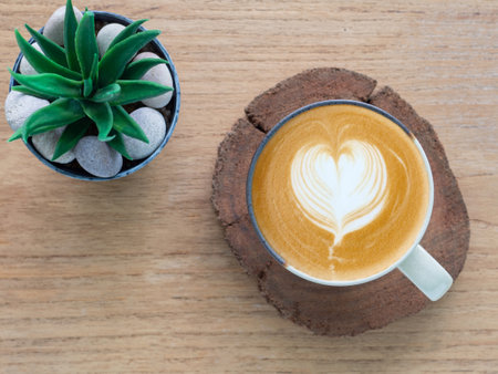 Top view of heart-shaped latte art coffee made with frothy milk in a ceramic mug with wooden saucer and small plant on wooden table. Cafe menu concept and banner.の写真素材