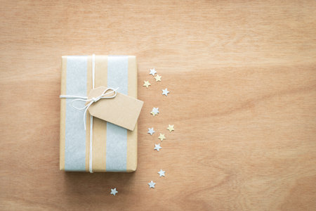 Gift box with brown tag surrounded by silver and gold paper star-shaped confetti on a wooden background. Top view, holiday concept, positive emotion, and birthday.の写真素材