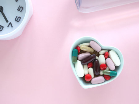 Top view, flat lay of blue heart-shaped bowl filled with supplements various colorful pills and capsules, clock on a pink wooden surface. Health, wellness, and care pharmacology concept.の写真素材