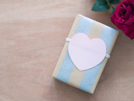 Flat lay, rectangular gift box, wrapped in kraft paper with silver stripes running vertically. A soft pink paper heart is centered on top of the gift and red rose on wooden background with copy space.の写真素材