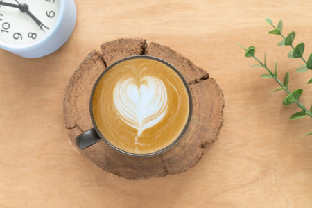 Concept for a banner, cafe, or coffee menu. A cappuccino latte art in the form of a heart in a dark brown ceramic cup, and a clock showing time 7:00, with green leaves on a rustic background.の写真素材