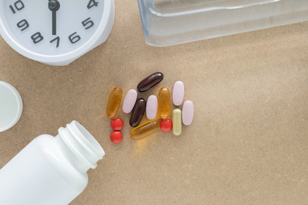 Top view, a group of scattered supplement pills and capsules and white plastic pill bottle, clock, bottle water on a brown background. Health, wellness, and care pharmacology concept.の写真素材