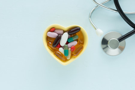 A yellow, heart-shaped bowl filled with various pills and capsules supplements is centered, and a stethoscope on a light blue wooden table. Top view, flat lay concept for health and care.の写真素材
