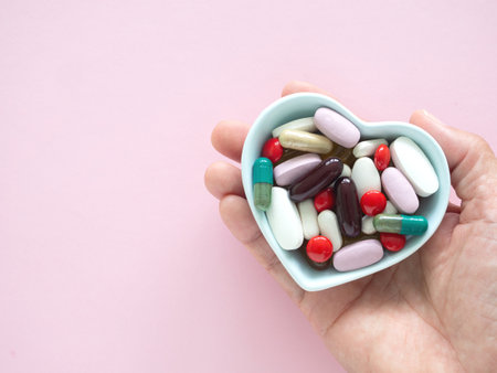 Hand holding a heart-shaped blue bowl filled with various colorful pills and capsules over pink background. Concept daily routines and medication management supplements.の写真素材
