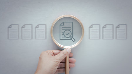 A hand holds a magnifying glass over a document icon that features a checkmark, suggests verification, approval, inspection, or selection over grey wooden background.の写真素材