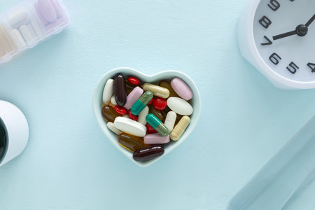 Top view, flat lay of blue heart-shaped bowl filled with supplements various colorful pills and capsules, clock on a light blue wooden surface. Health, wellness, and care pharmacology concept.の写真素材