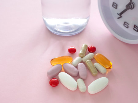 A collection of supplements of various pills and capsules scattered and clock, water bottle on a light pink background. Close-up with copy space. Health supplement concept.の写真素材