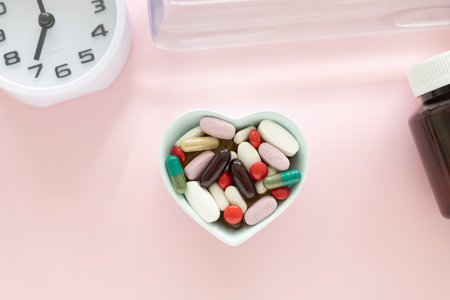 Light blue heart shaped bowl with various colored dietary supplement pills and clock on pink background. Health supplement concept. Top view with copy space.の写真素材