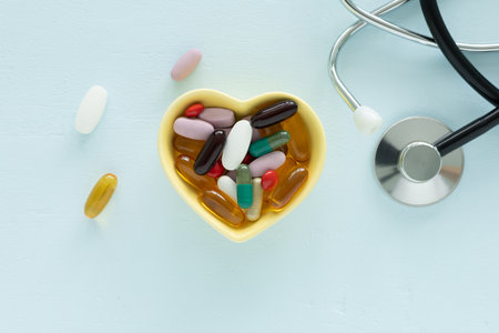 A yellow, heart-shaped bowl filled with various pills and capsules supplements is centered, and a stethoscope on a light blue wooden table. Top view, flat lay concept for health and care.の写真素材