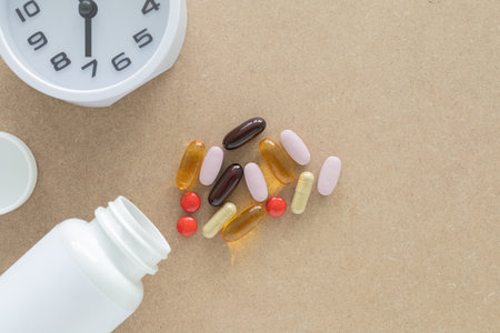 Top view, a group of scattered supplement pills and capsules and white plastic pill bottle, clock, on a brown background. Health, wellness, and care pharmacology concept.の写真素材