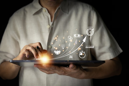 Business growth and strategy concept. Man holding a tablet displaying an up arrow with icons representing money, ideas, team work, and a target for 2026. Digital transformation and future planning.の写真素材