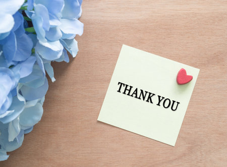 A yellow sticky note with "THANK YOU" written on it is placed on a wooden desk with a small red heart with a blue bouquet of flowers. symbolizing gratitude, appreciation, and special moments.の写真素材