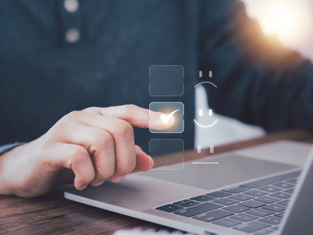 Online survey and feedback concept. Hand checking a box with a glowing checkmark on a virtual interface, next to emoji options, above a laptop. Customer satisfaction. Digital feedback form completion.の写真素材