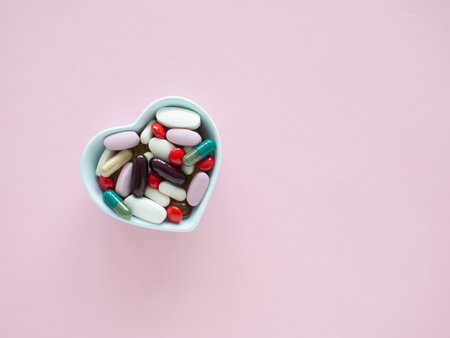 Top view, flat lay of blue heart-shaped bowl filled with a colorful assortment of various supplements on a pink wooden background, health, wellness, and care pharmacology concept.の写真素材