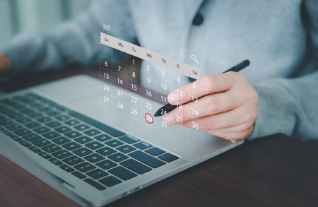 A professional's hand is using a stylus, pointing to a virtual calendar projected over a laptop, with a specific date circled. Concept for efficient digital planning, time management, and deadlines.の写真素材