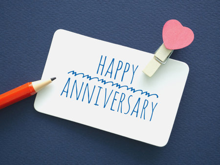 Top view a small card with the words "Happy Anniversary" written in a friendly, blue font. A pink heart is attached with a clothespin, a red pencil over dark blue background. Love and relationships.の写真素材