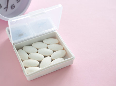 The white pill box is open and contains white plastic pill organizer and clock on pink table. Health, wellness, and care pharmacology concept. Close up.の写真素材