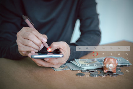 Person using a smartphone to manage savings and budgets, with a calendar, piggy bank, and cash on the desk. Digital banking, wealth management, and retirement goals for future investment.の写真素材