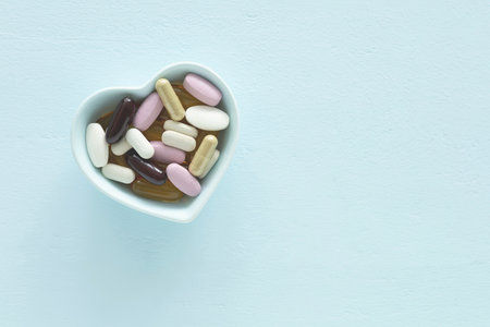 Top view, flat lay of blue heart-shaped bowl filled with supplements various colorful pills and capsules on a light blue wooden surface. health, wellness, and care pharmacology concept.の写真素材