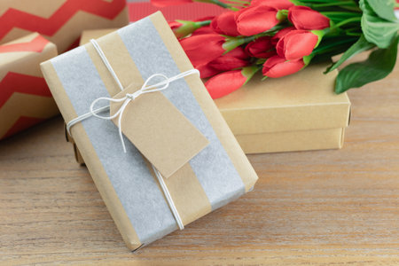 A wrapped gift box with kraft paper and silver stripes, tied with a simple white string and blank tag on a wooden table. and a vibrant bouquet of red tulips.の写真素材