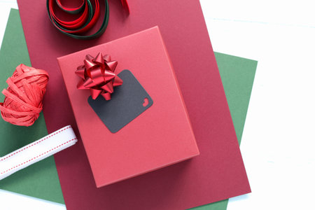 Top view flat lay of Christmas gift wrapping. A red box with a bow and blank black chalkboard tag for mockup. Festive holiday supplies, paper, and ribbons on a white wood background.の写真素材