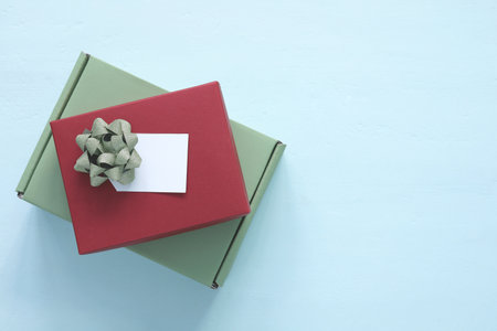 Stacked red and green gift boxes with gold glitter bow and blank white tag isolated on blue wooden background. Top view of present for Christmas or birthday celebration concept with copy space.の写真素材