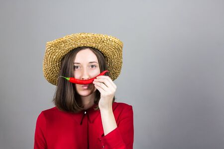 Girl in red with chili pepper as mustache. Young woman in traditional hat and red shirt posing with pepper holding it as mustache in front of studio backgroundの写真素材