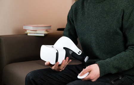 Holding virtual reality headset and remote control. Person on sofa in living room with vr glasses...