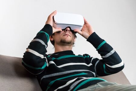 Young male person sits on sofa in vr glasses, adjusting them comfortable with hands