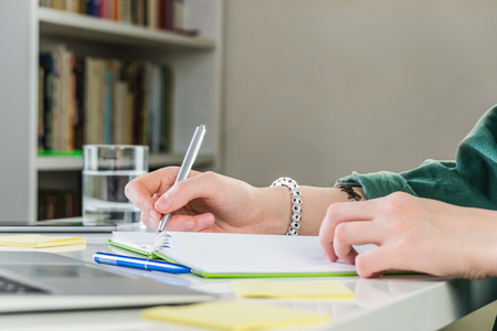 Taking notes in copybook at home working place. Female person hand writes in notebook sitting at the table indoors in front of laptopの写真素材