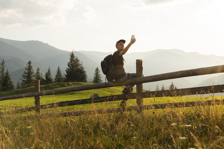 male hiker sits on fence in ukrainian carpathian mountains and communicates by mobile phoneの写真素材