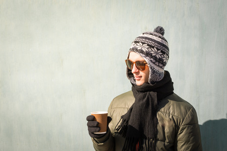 Young man in winter clothes holds cup of coffee. Handsome teenage male person in sunglasses and...