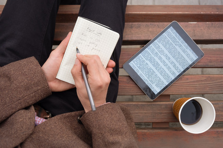 Shot from above of stylish male person taking notes and working sitting on wooden bench outdoors....