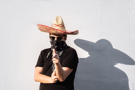 Young male person dressed up in traditional sombrero, bandana and sunglasses. Mexican festive or halloween concept of man posing as bandit or western style gangsterの写真素材