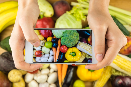 Taking photo of fresh organic fruit and vegetables on rustic wood table, top view. Close-up of female hands photographing flat lay of locally grown natural vegan foodの写真素材