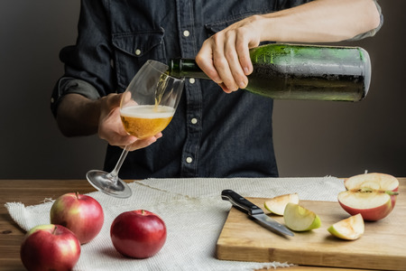 Male hands pouring premium cidre in wine glass above rustic wood table. Man pours a glass of vintage apple wine out of ice cold bottleの写真素材