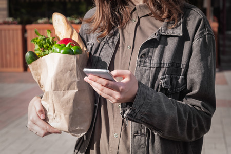 Checking shopping list in smarphone while shopping for food. Attractive young woman in casual style clothes holding Recyclable paper bag of groceries bought from local vegetable and grocery store or market.の写真素材