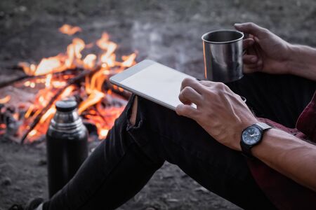 Working with technology in unconventional place concept. Using a tablet computer to work remotely at a hiking trip next to camping fire.の写真素材