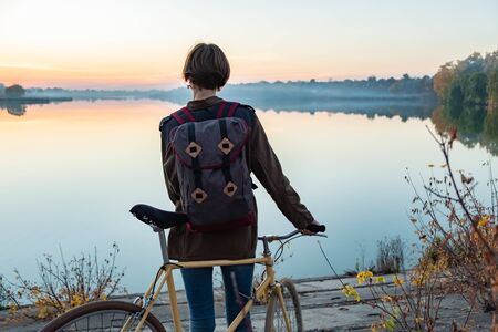 Female cyclist enjoying beautiful blue hour scene by the lake. Woman stands with bike and looks at beautiful lake and sunsetの写真素材