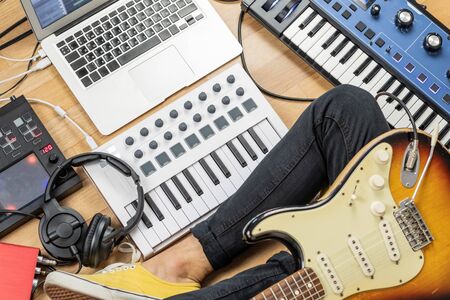 Male guitarist with electric guitar at modern home studio or rehearsal room. Young man producing music with electronic effects processors, synthesizer and laptopの写真素材