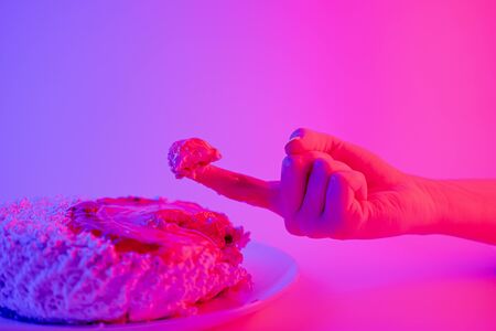 Being a sweet tooth concept: finger with a piece of cake. Partying, celebration or being impatient concept: sticky finger and a cake against vibrant neon backgroundの写真素材