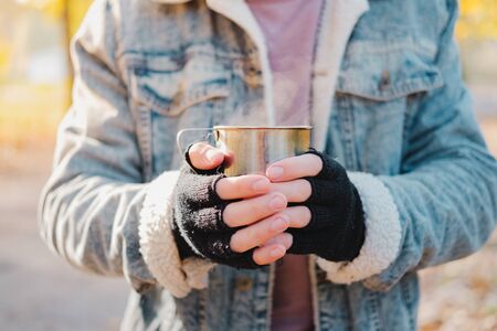 Man in mittens keeps warm holding a cup of hot drink. Chilly morning outdoors scene: human hands...