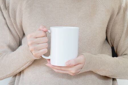 Woman in warm sweater is holding white mug in hands